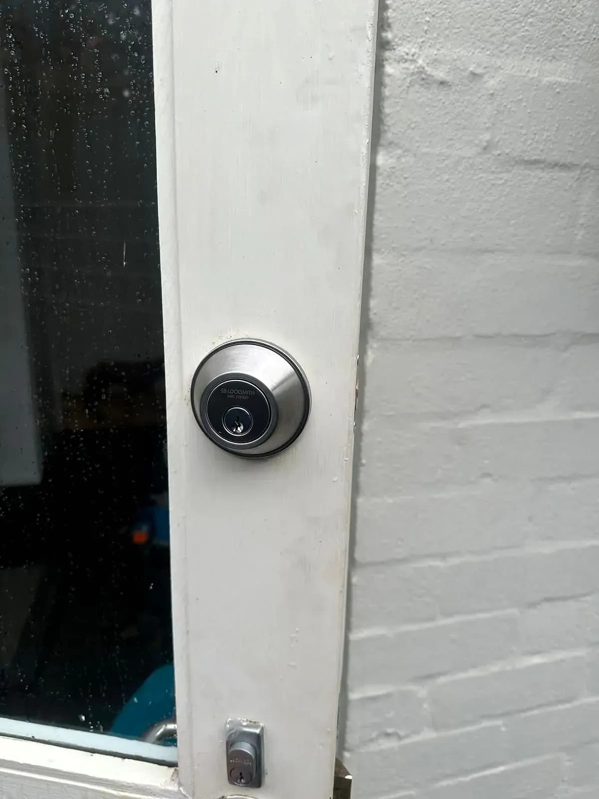 A deadlock on a white timber door beside a window, its cylinder engraved with "SB Locksmith" and the phone number