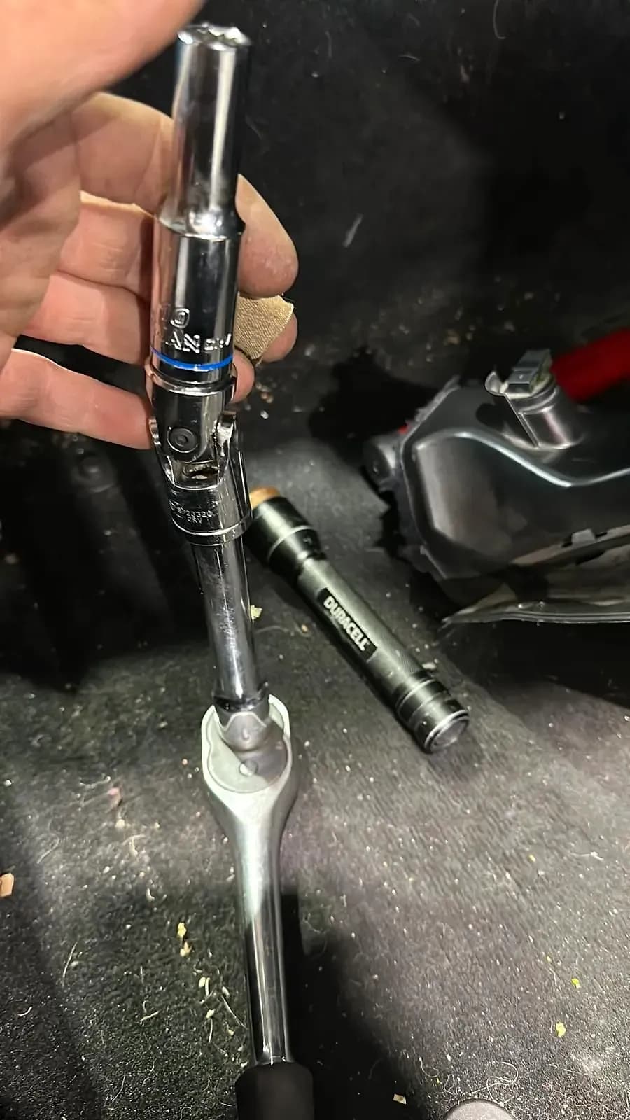 A hand holding a ratchet with a long socket on a universal joint over a car’s footwell, a torch on the carpet beside a removed trim panel
