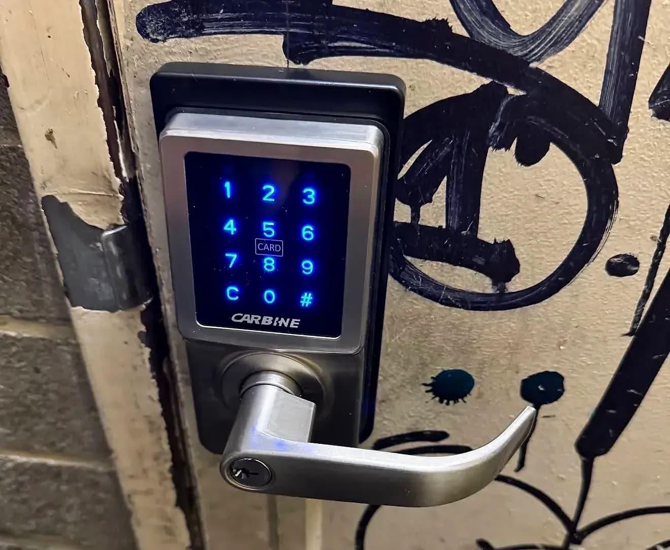 A Carbine keypad and card lever lock lit blue on a door set in a graffitied laneway wall
