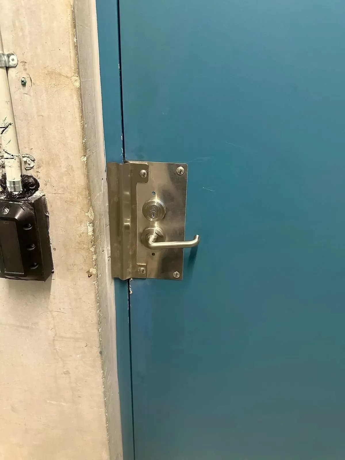 A stainless lock guard plate with a lever handle and a key cylinder on a teal steel door in a building service area