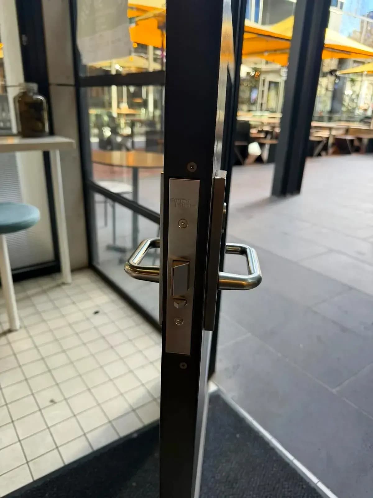 A dormakaba mortice lock in the edge of a glass café door, with stainless D-handles either side and the street beyond