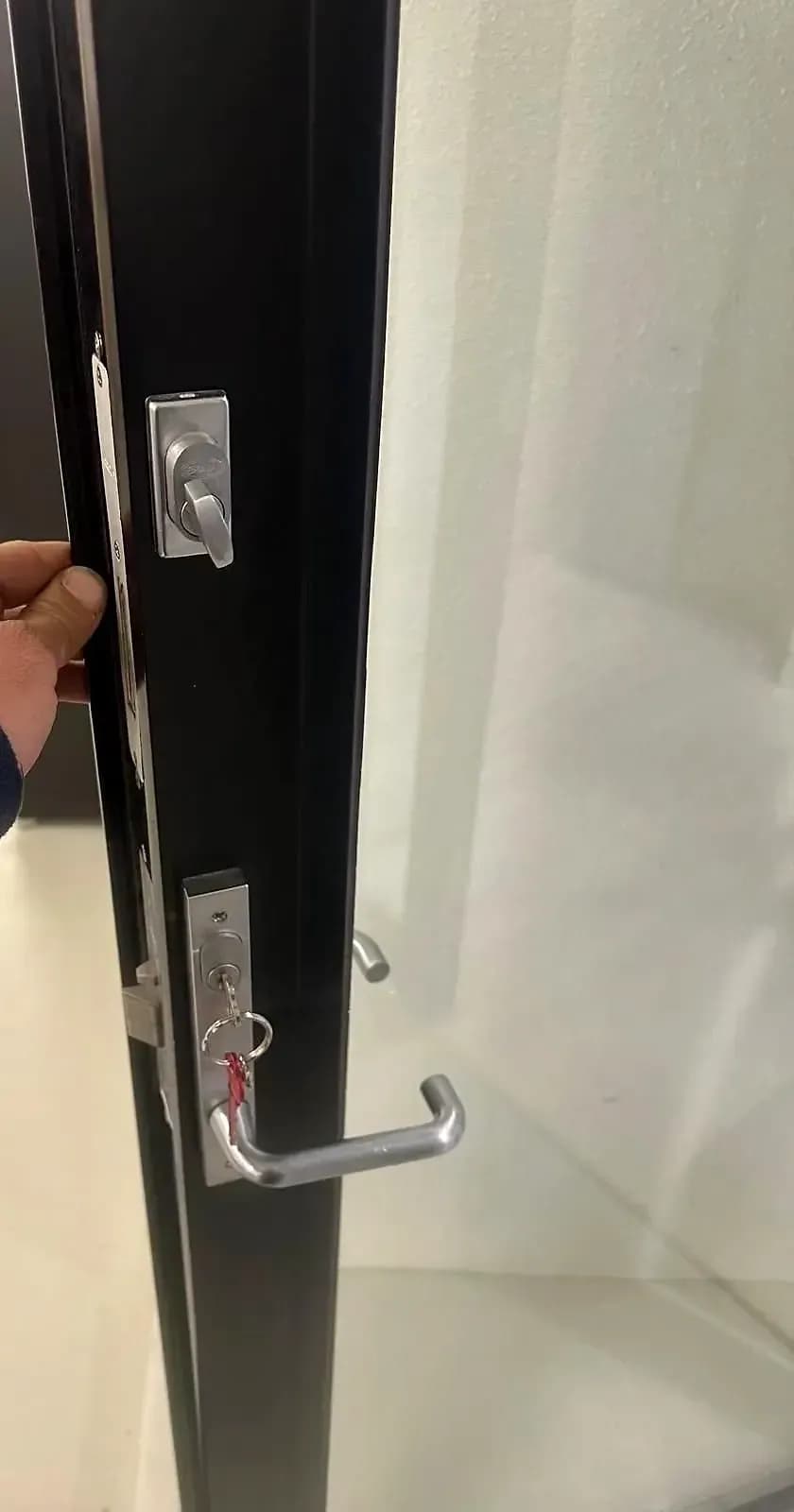 A hand holding open a black-framed glass door fitted with a thumb turn above and a keyed lever lock below, the new keys in the cylinder