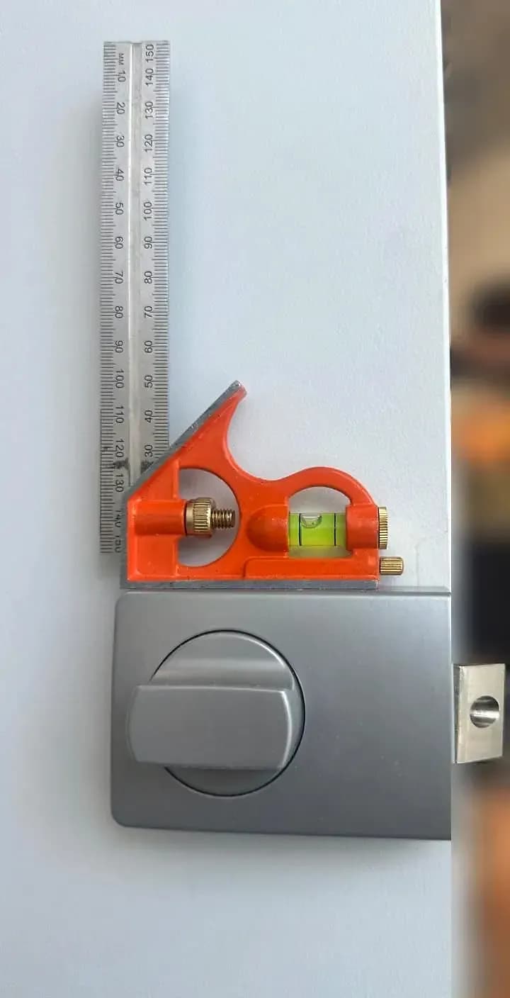 A combination square resting on a newly fitted deadlock, its bubble level, on the face of a white door near its edge