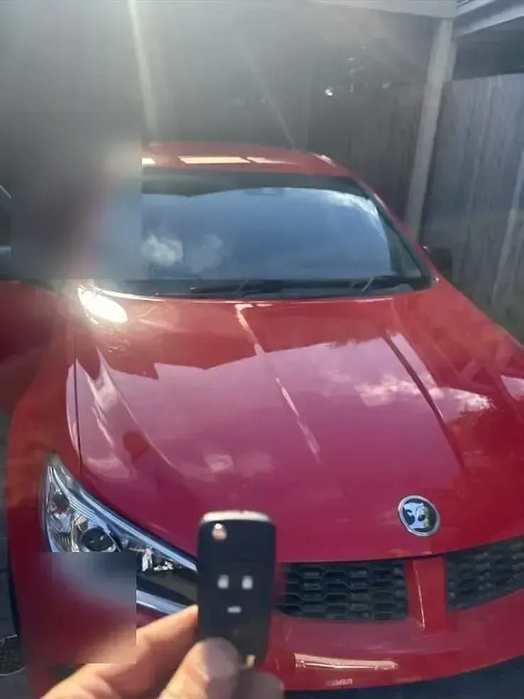 A Holden flip key held up in front of a red Holden Commodore under a carport