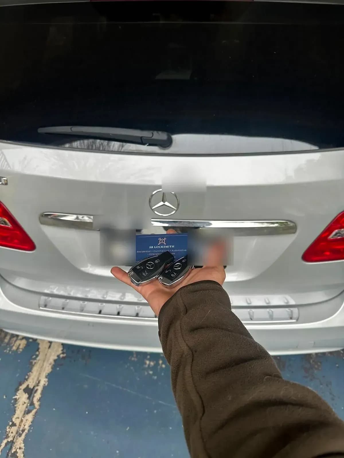 Two Mercedes-Benz smart keys and an SB Locksmith business card held out behind a silver Mercedes B-Class, its number plate blurred