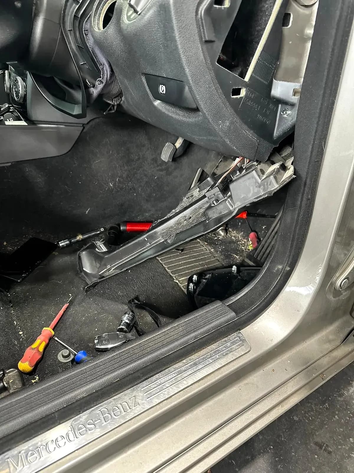 The driver’s footwell of a Mercedes-Benz with the lower dash trim removed, screwdrivers and sockets on the carpet and the smart key on the sill