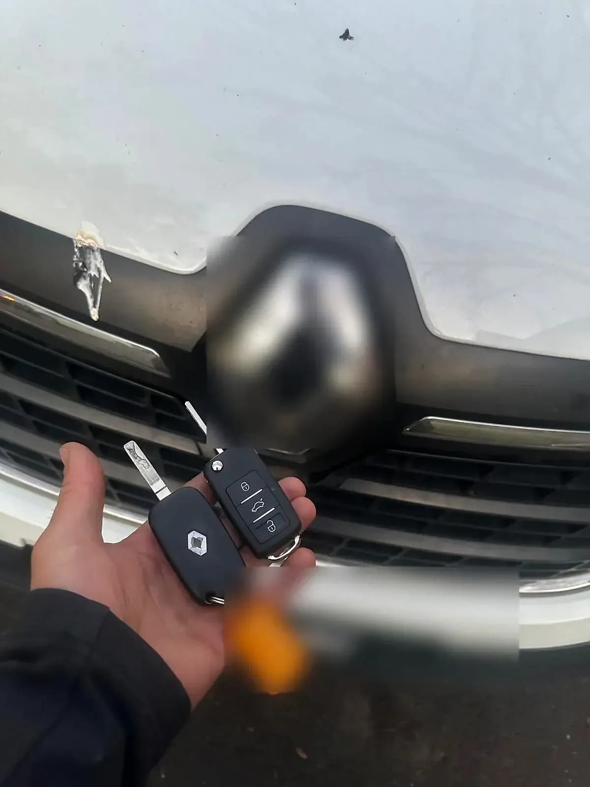 A Renault flip key and a new flip key held up in front of the grille of a white Renault