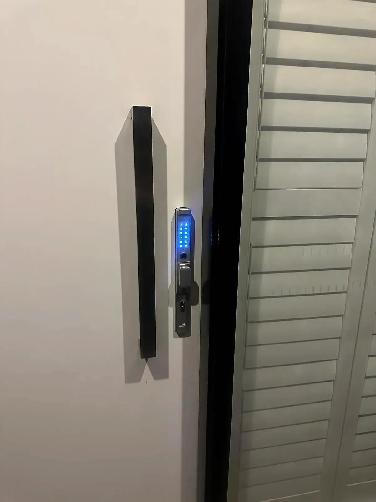 A slim keypad smart lock lit blue on a black-framed sliding door, beside a long pull handle