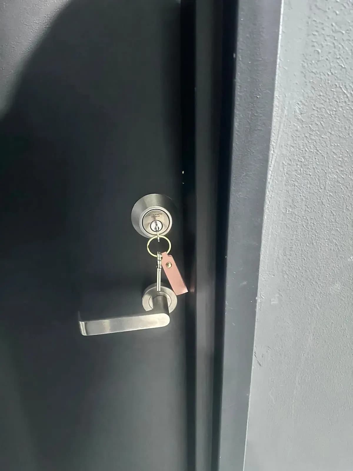 A deadbolt on a black door with "SB Locksmith" engraved on the cylinder, the new keys hanging from it on a leather tag, a lever handle below