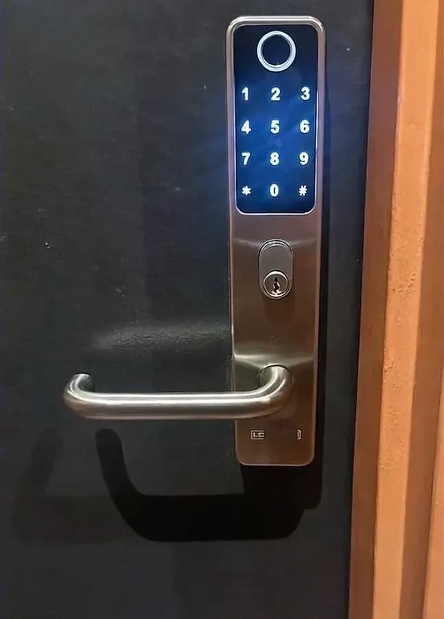 A stainless keypad and fingerprint lock with a lever handle, lit blue, retrofitted to a dark door